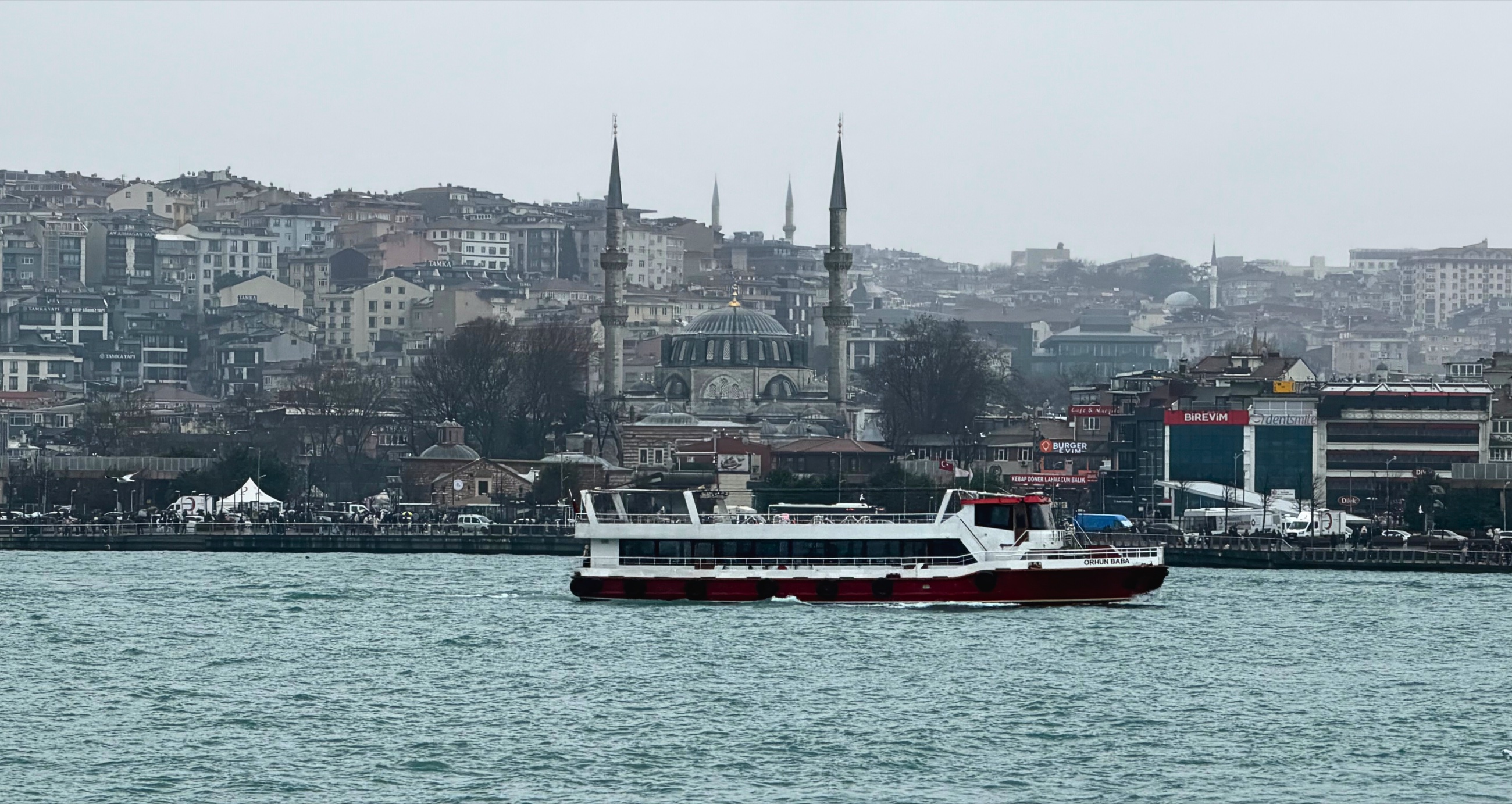 A passenger ferry crossing the water in Istanbul with mosques and hillside buildings behind it.