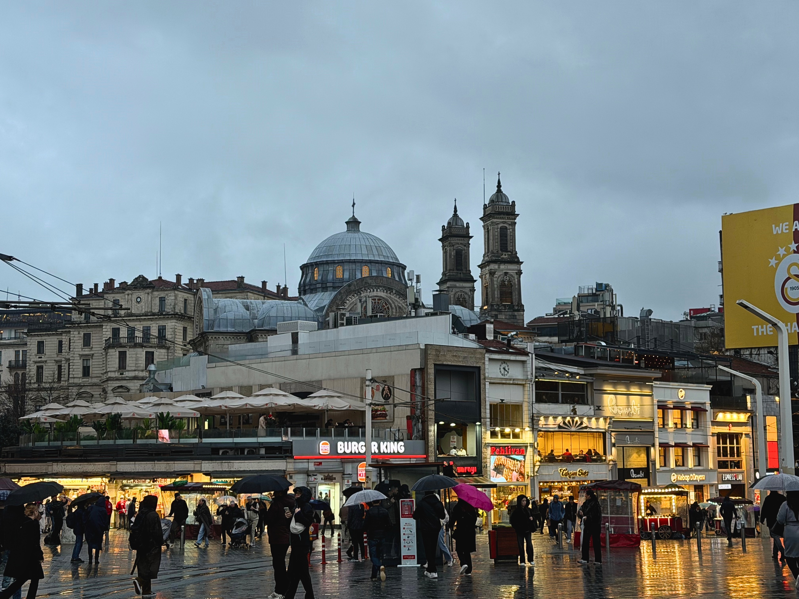 A rainy scene at Taksim with pedestrians, umbrellas, storefronts, and a domed church in the background.