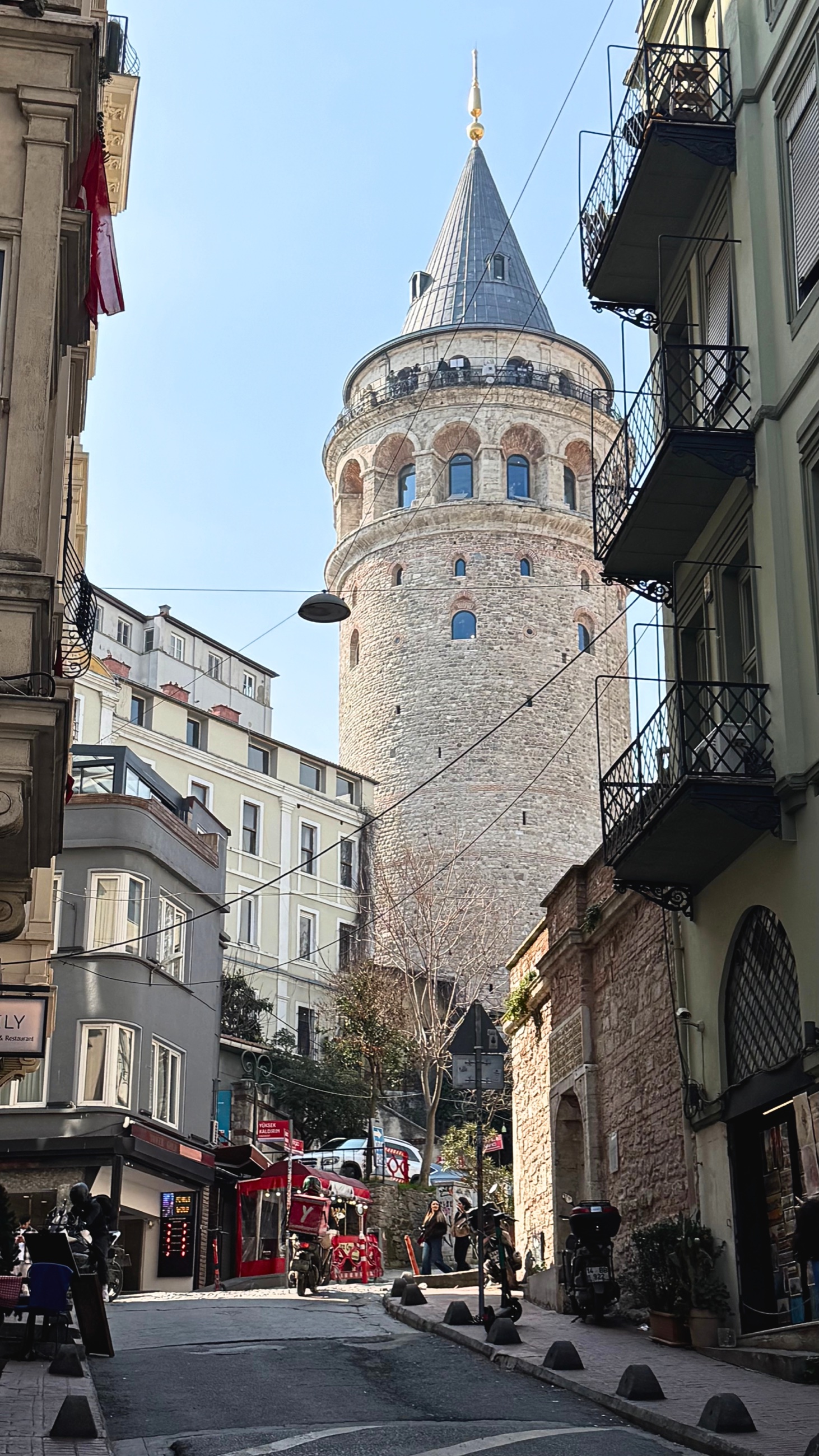Galata Tower rising above a narrow street lined with buildings, balconies, and pedestrians.