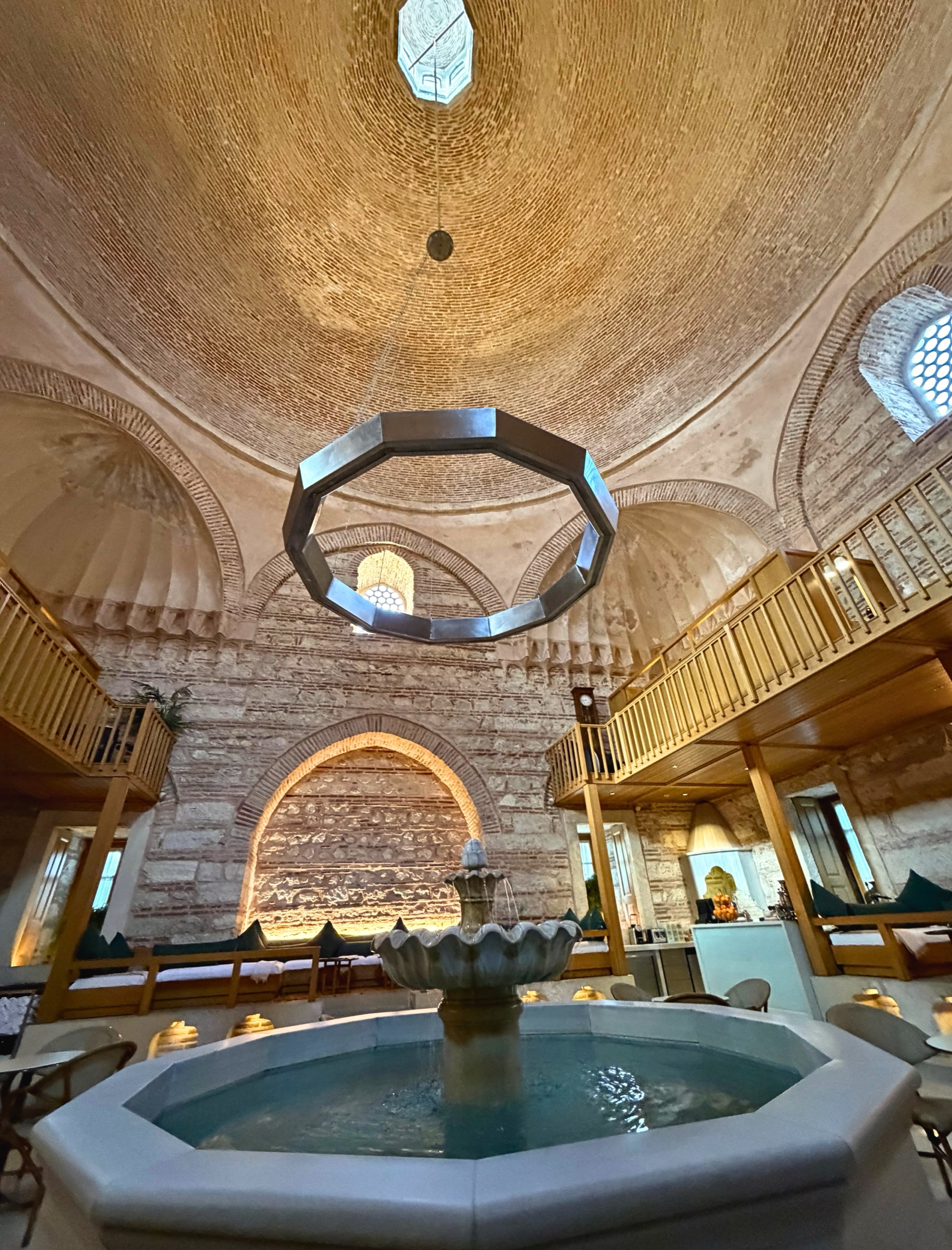The interior of a historic hamam with a domed brick ceiling, fountain, warm lighting, and stone walls.