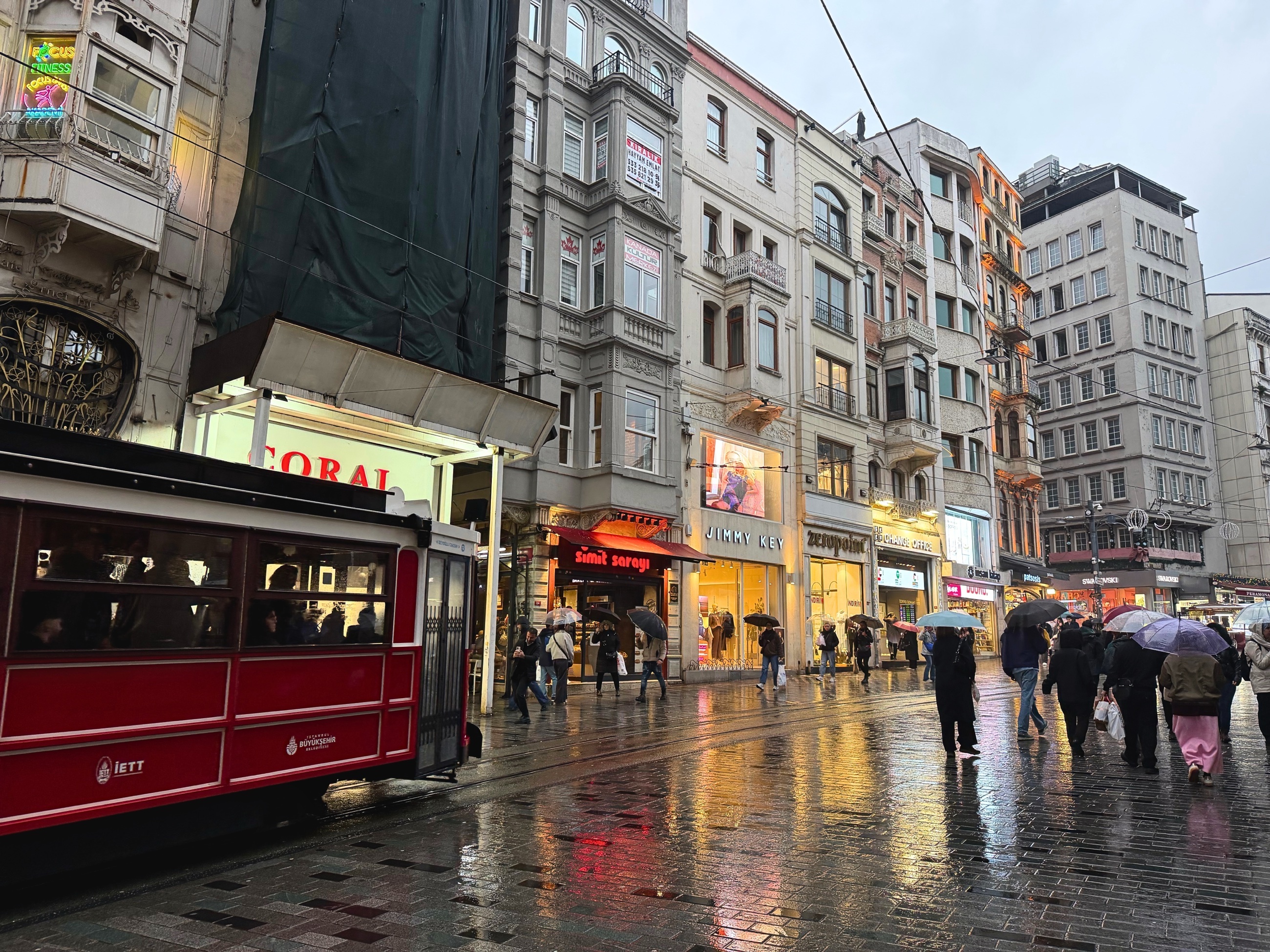 The red tram on Istiklal Avenue during evening rain, with wet pavement, shopfront light, and pedestrians.