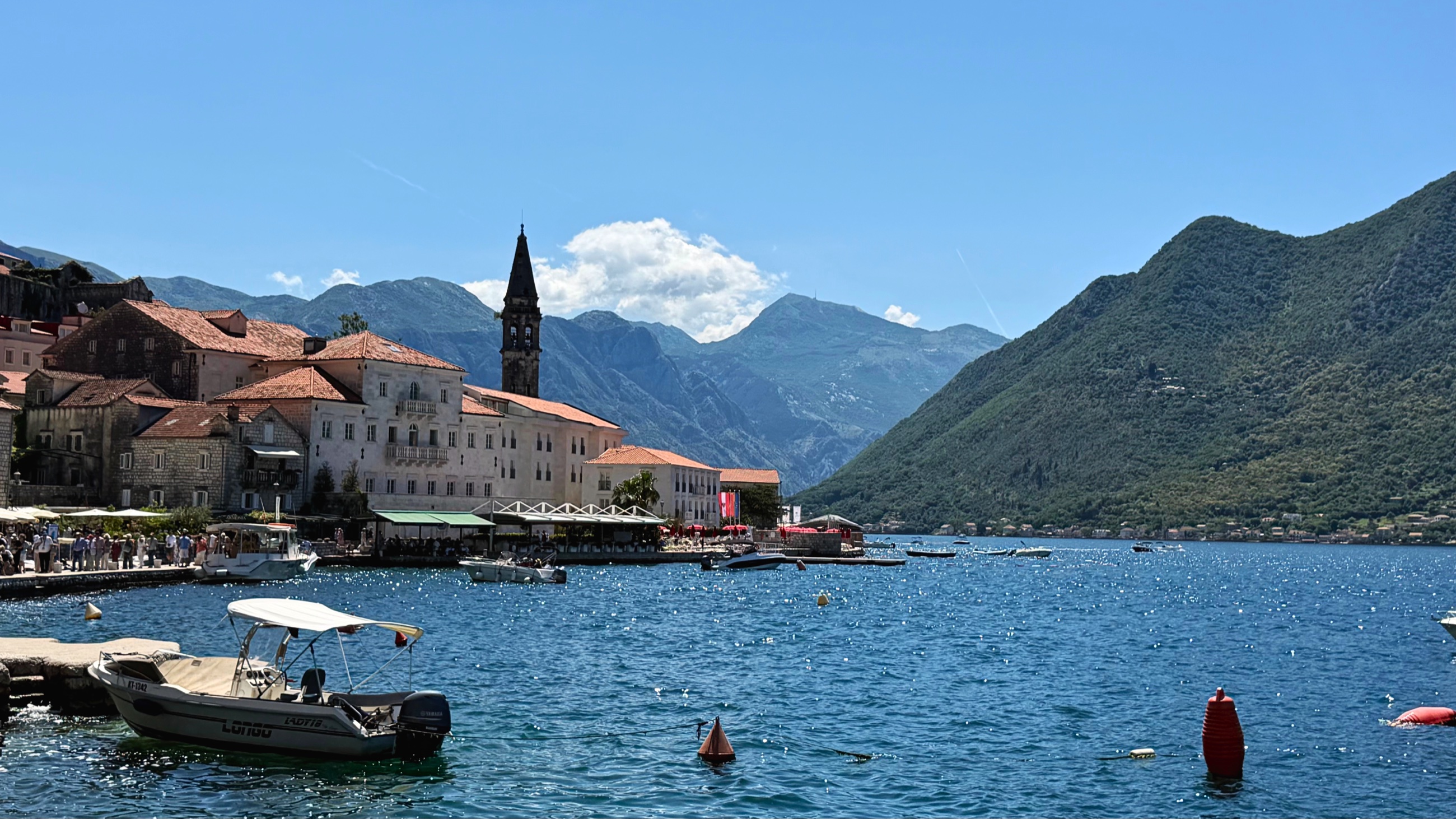 Perast from the waterline, with stone houses, church towers, boats, and mountains along the bay.