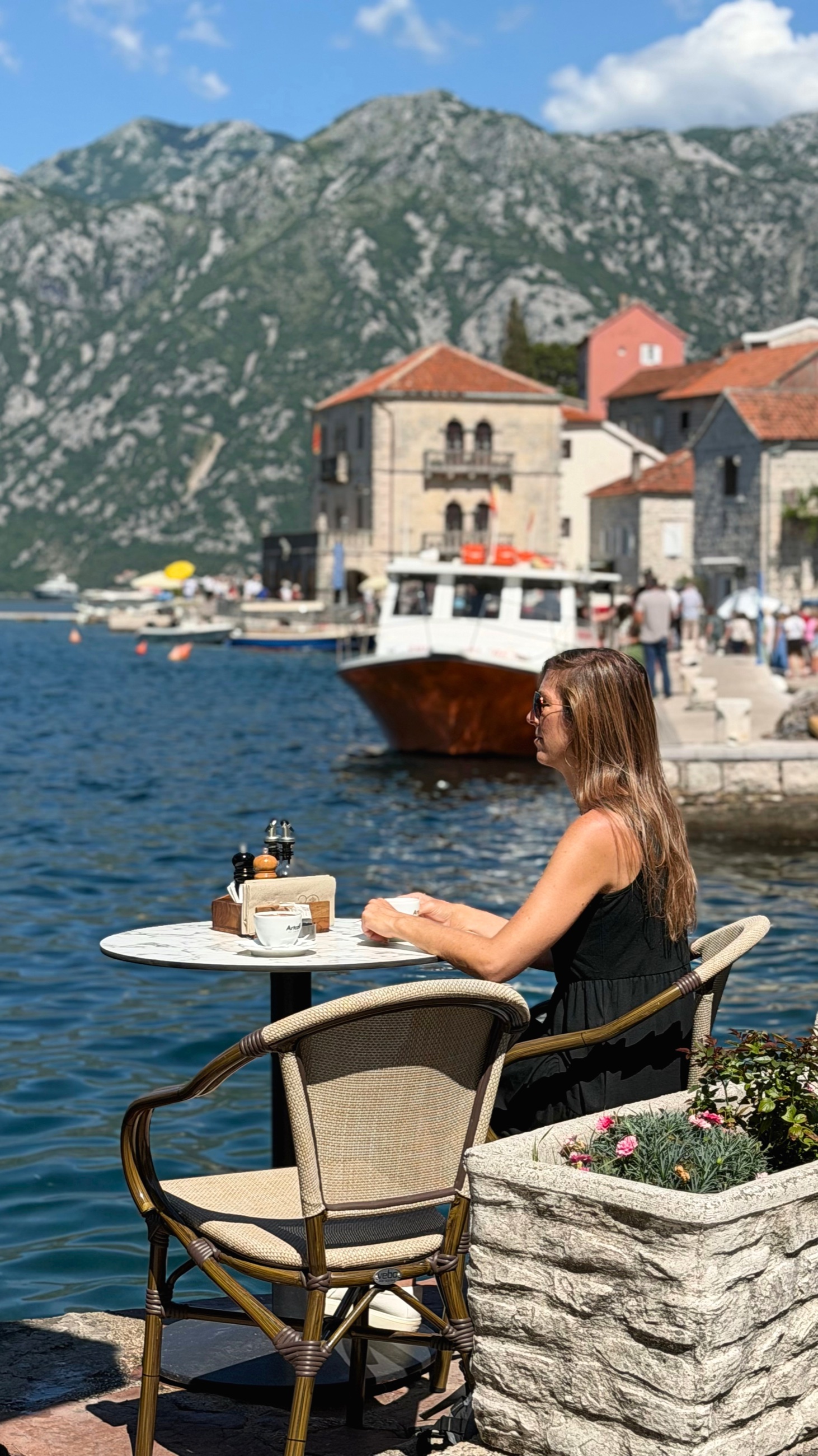 Coffee by the water in Perast, with Stephanie seated beside the bay and mountains behind.