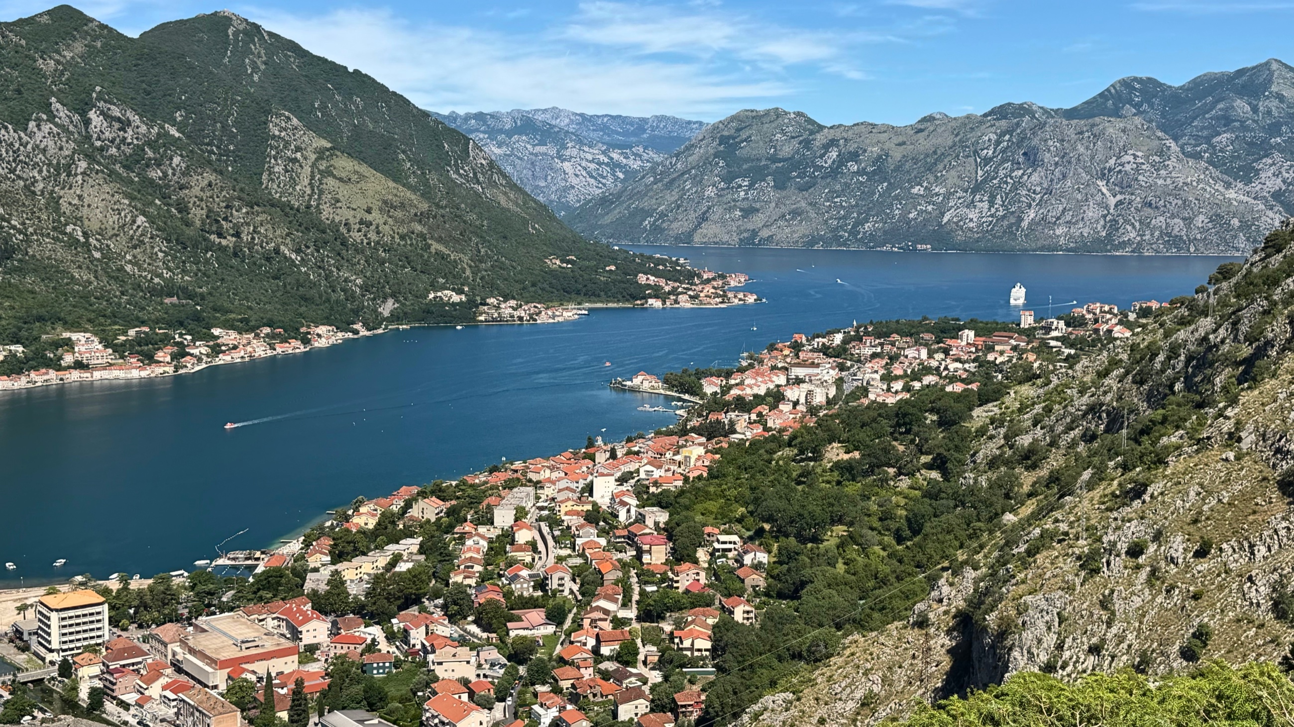 The Bay of Kotor from above, with Kotor and Dobrota merged into the wider shape of the bay, red roofs, blue water, steep mountains, and one cruise ship