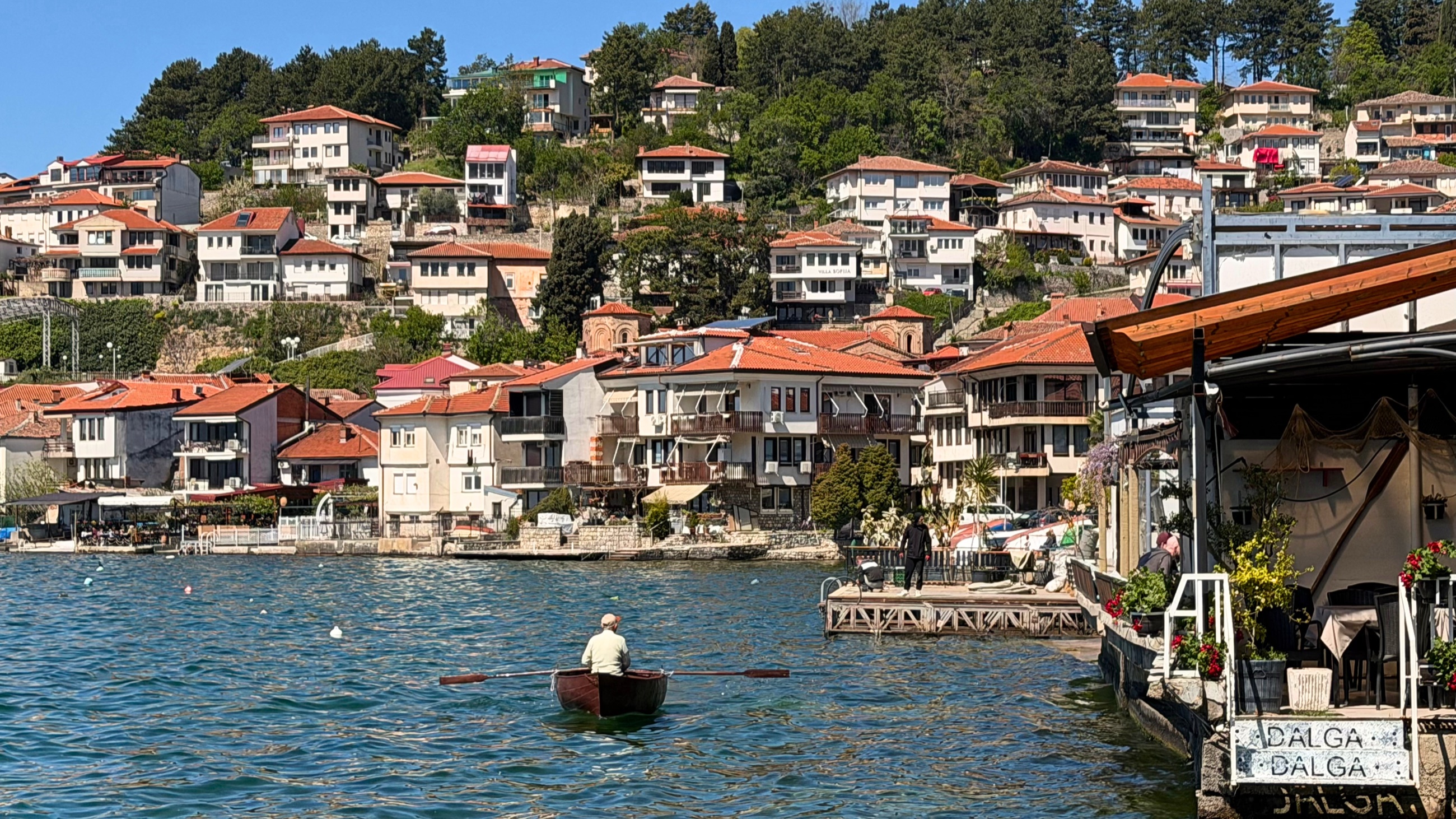A rowboat crossing Lake Ohrid below hillside homes, terracotta rooftops, lakefront terraces, and old churches.