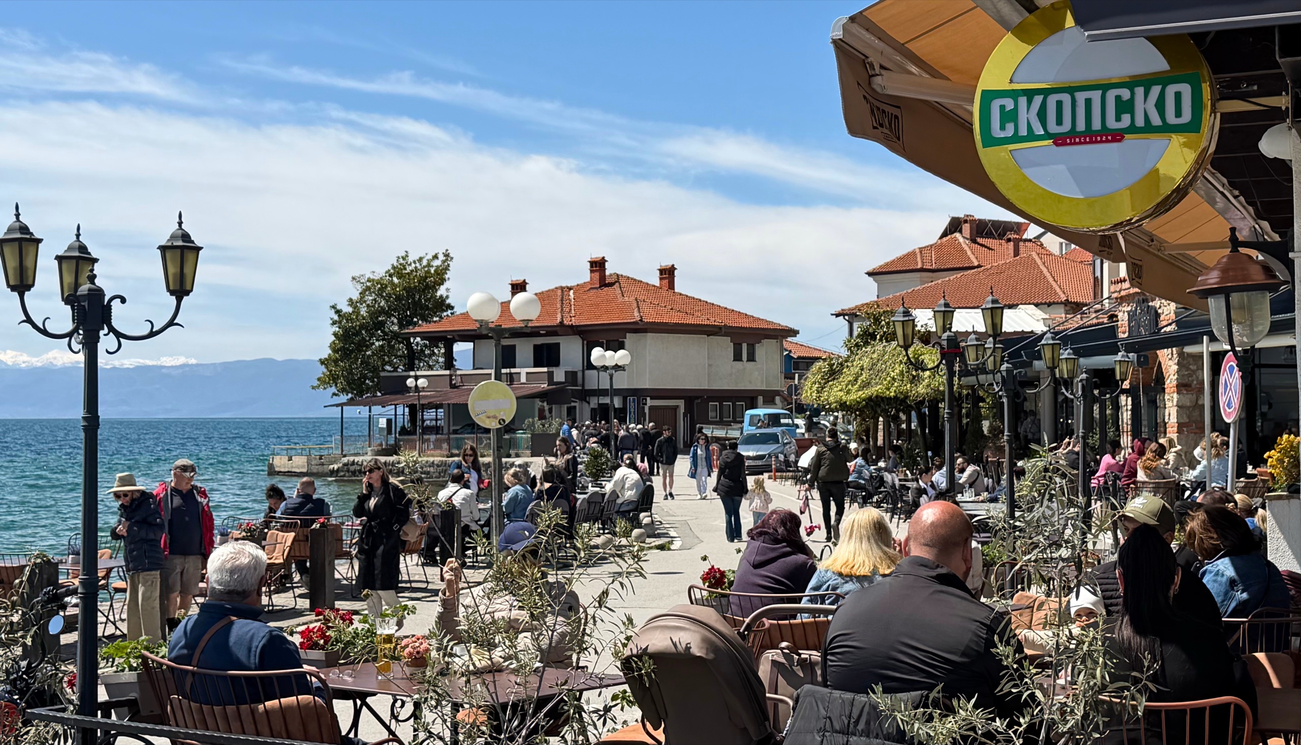 A busy sunny waterfront lane in Ohrid with cafés, people, lake views, and old town buildings.