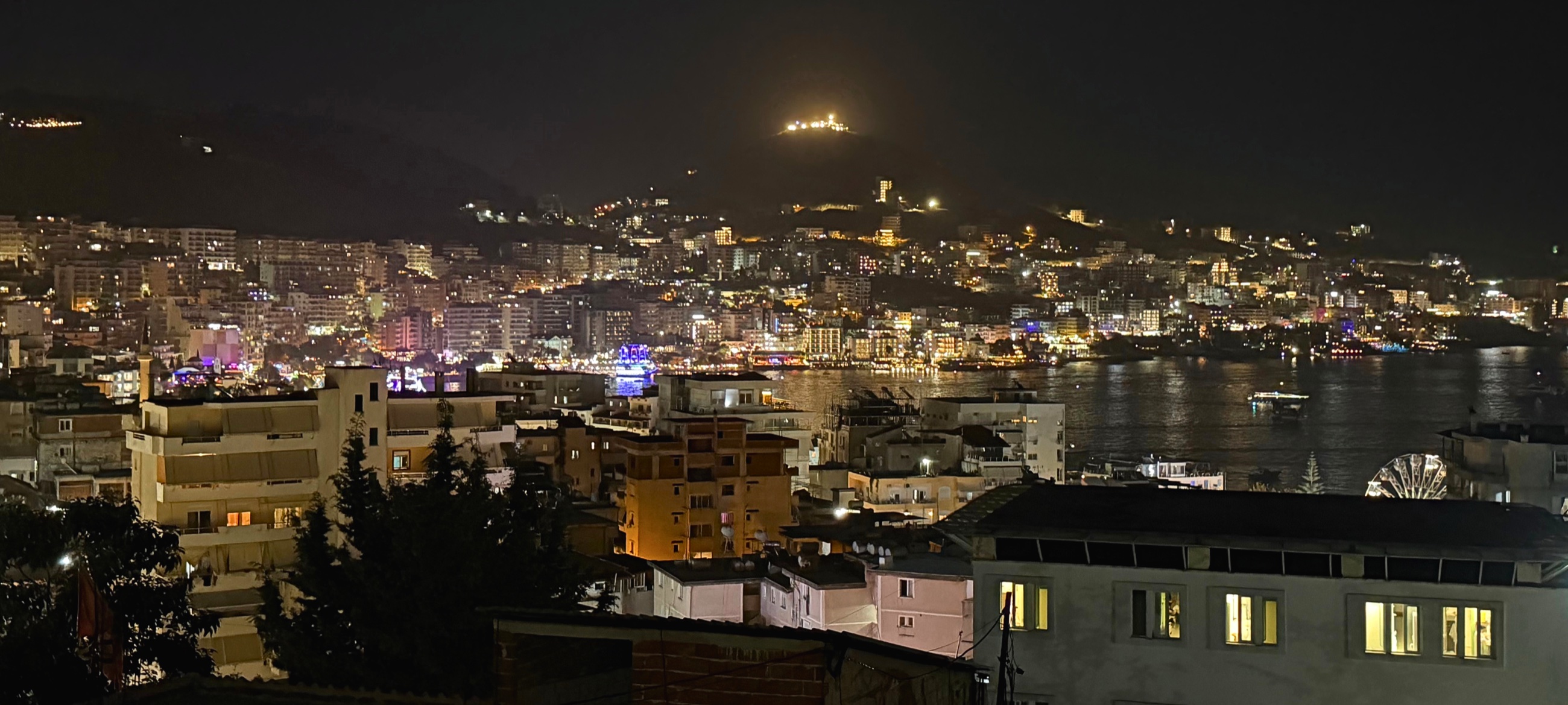 Sarandë illuminated at night along the Ionian coast, viewed from an apartment above the city.