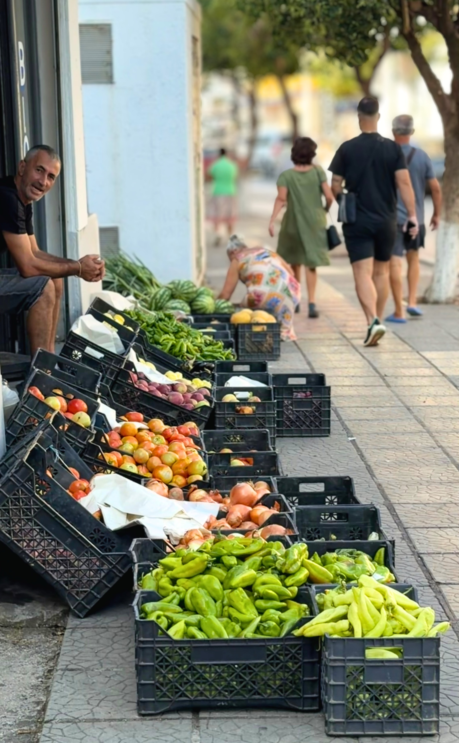 Crates of peppers, tomatoes, onions, melons, and other produce displayed along a Sarandë sidewalk market.