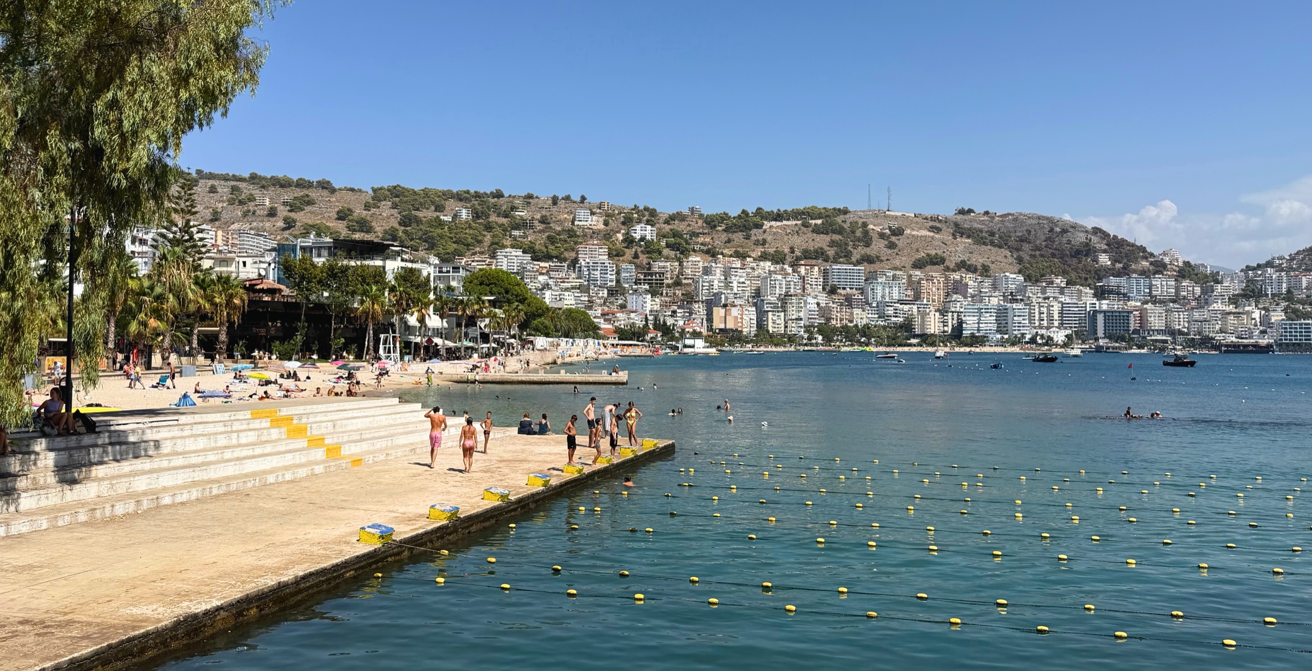 People swimming and relaxing along the Sarandë shoreline with the hillside city rising behind the bay.