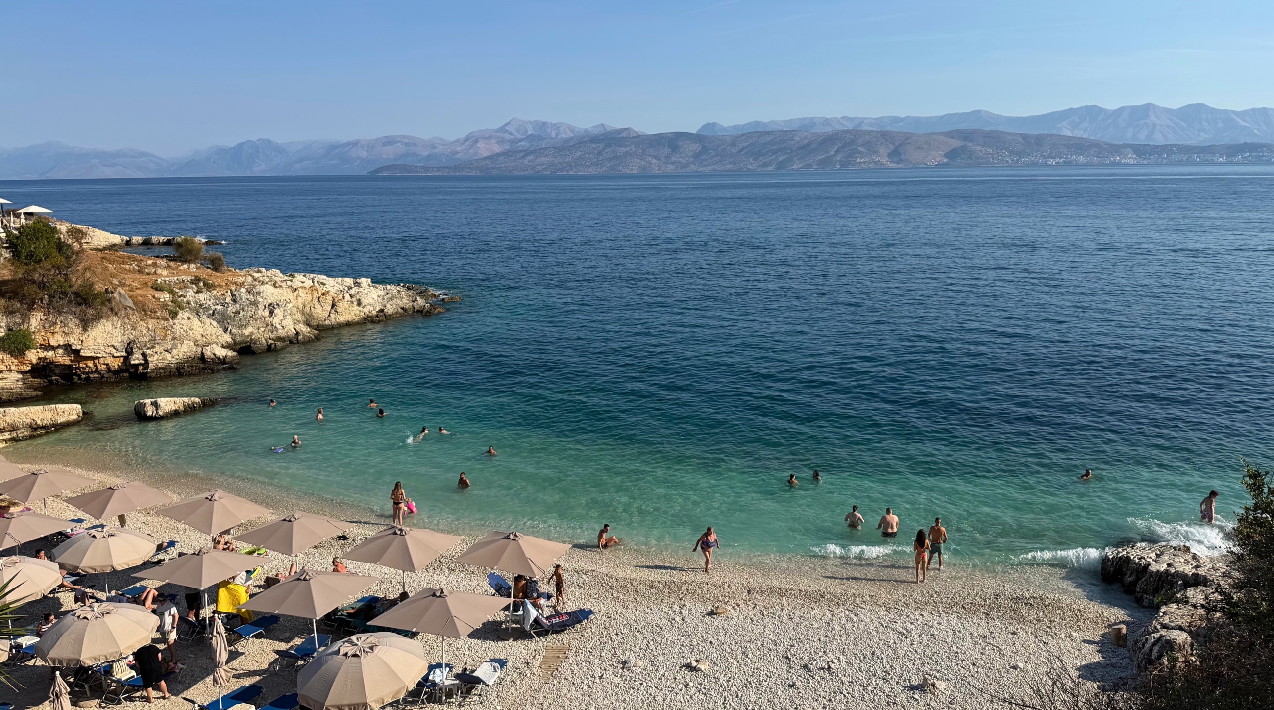 Bataria Beach in Kassiopi, Corfu, with swimmers in clear Ionian water and the Albanian coast across the channel.