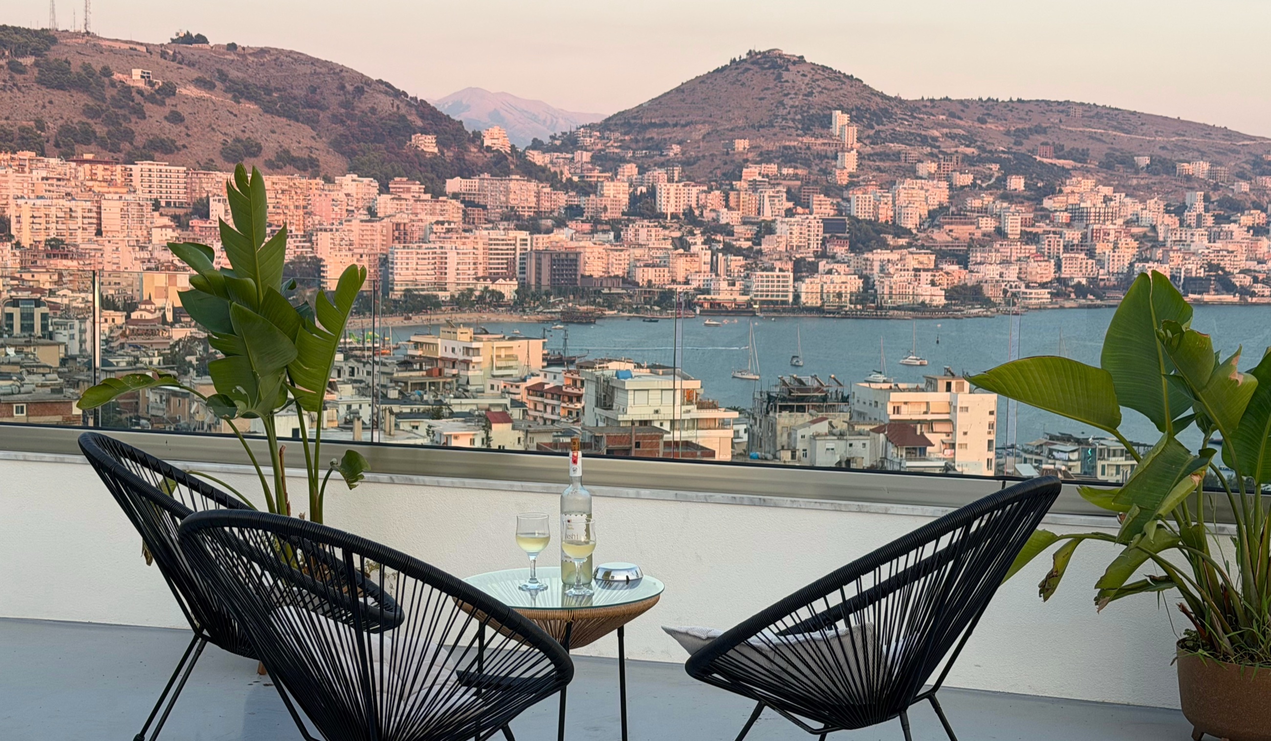 Two chairs, wine, and a view across Sarandë from a rooftop terrace at sunset.