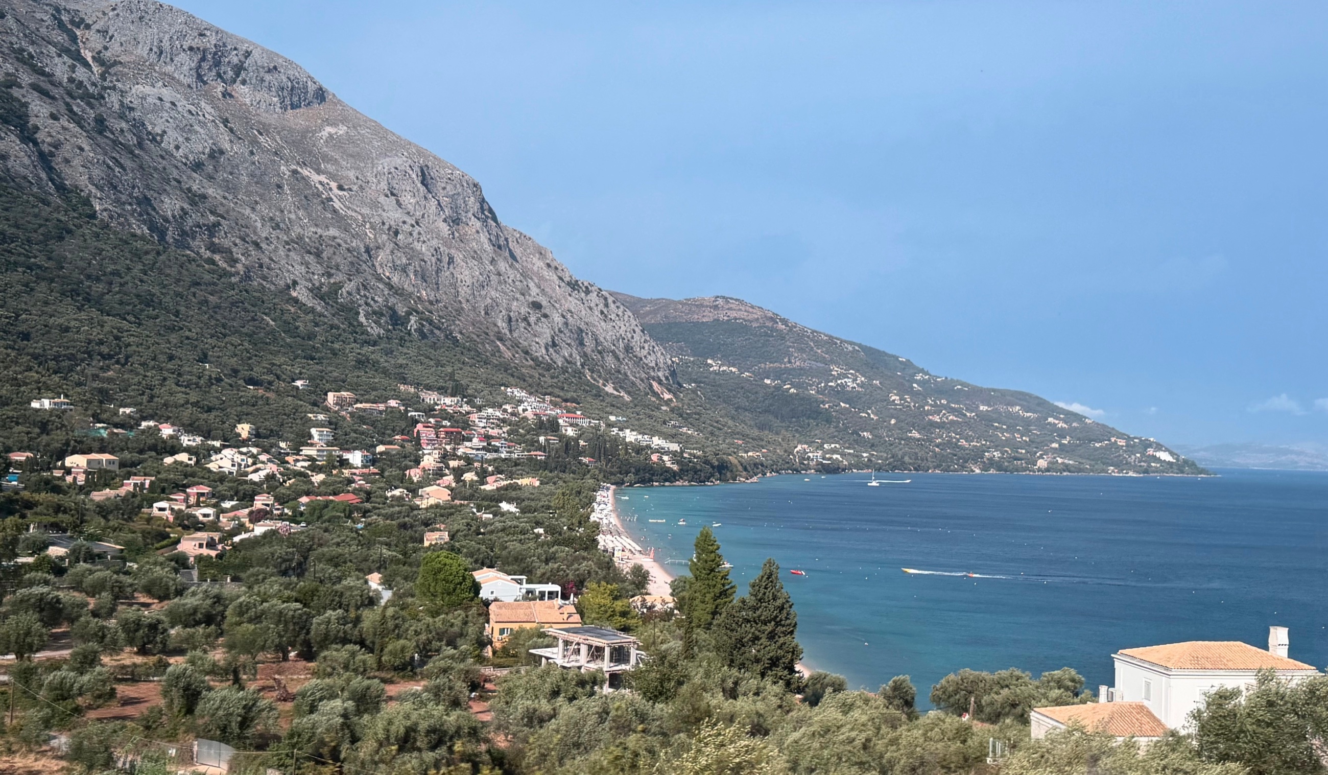 The steep eastern coast of Corfu on the road north toward Kassiopi, with villages beneath the mountain and the Ionian Sea alongside.