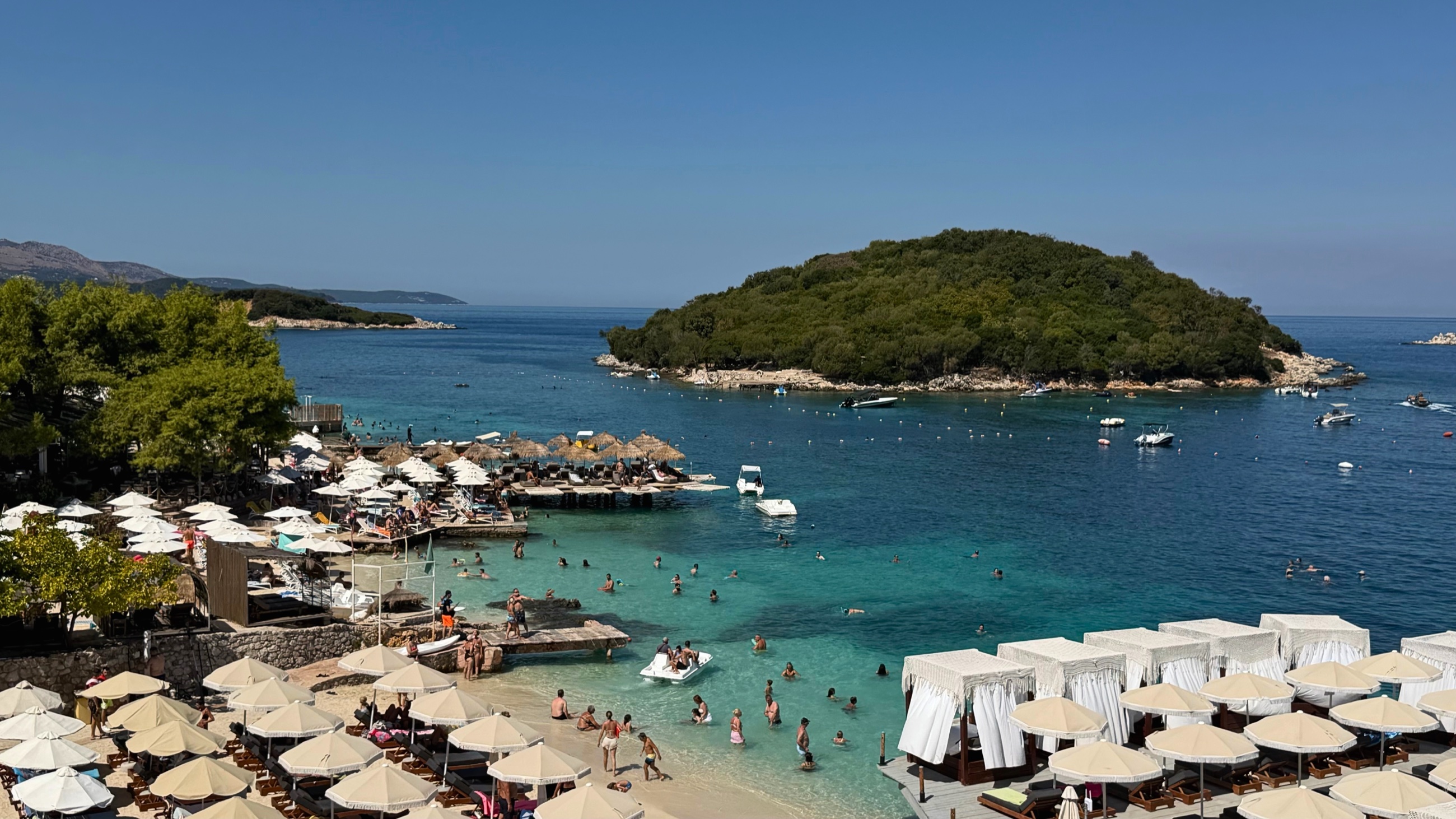 A busy turquoise bay at Ksamil with swimmers, loungers, umbrellas, and a small island offshore.