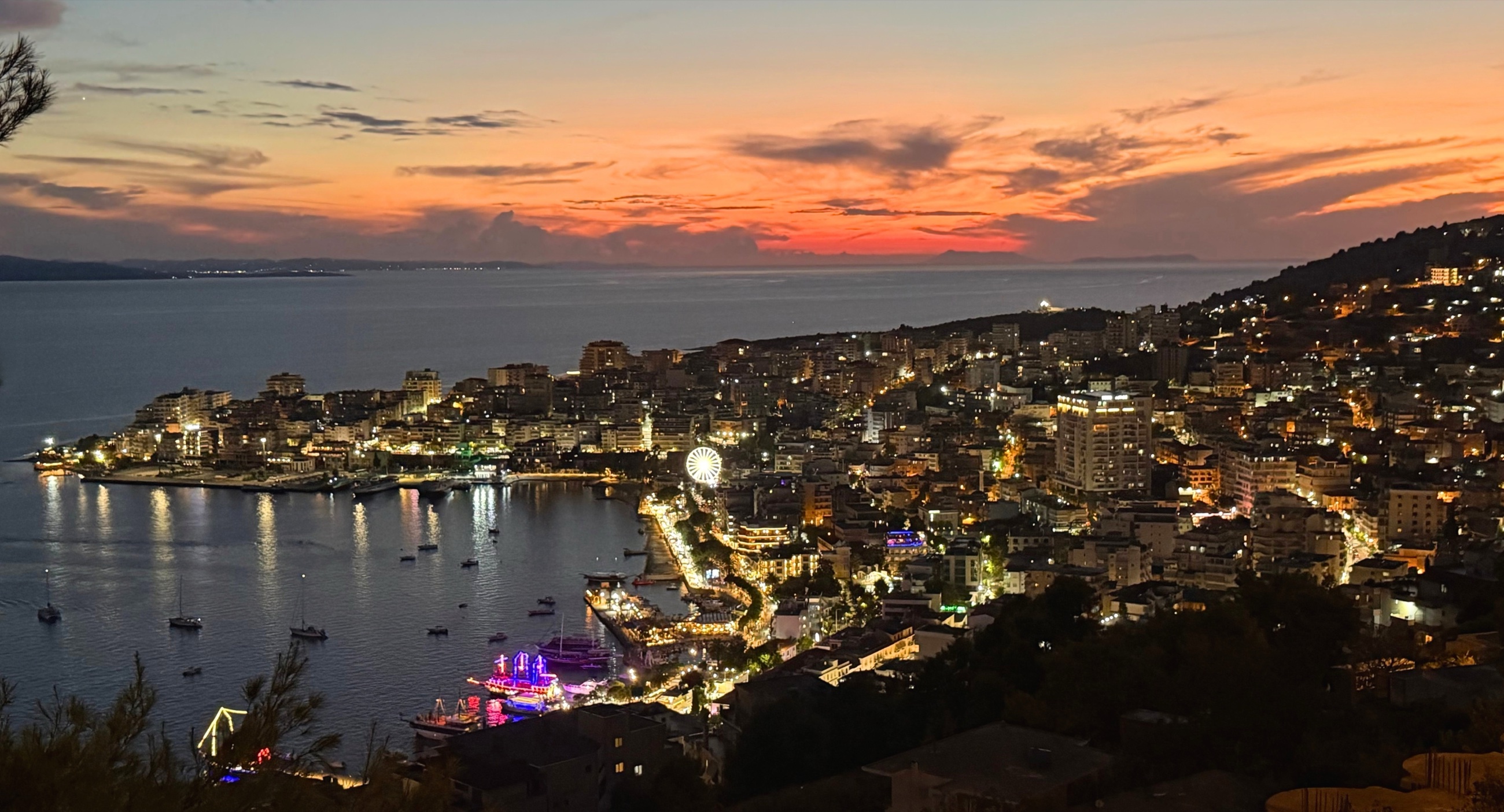 Sarandë Bay at dusk, with orange sky and lights along the waterfront.