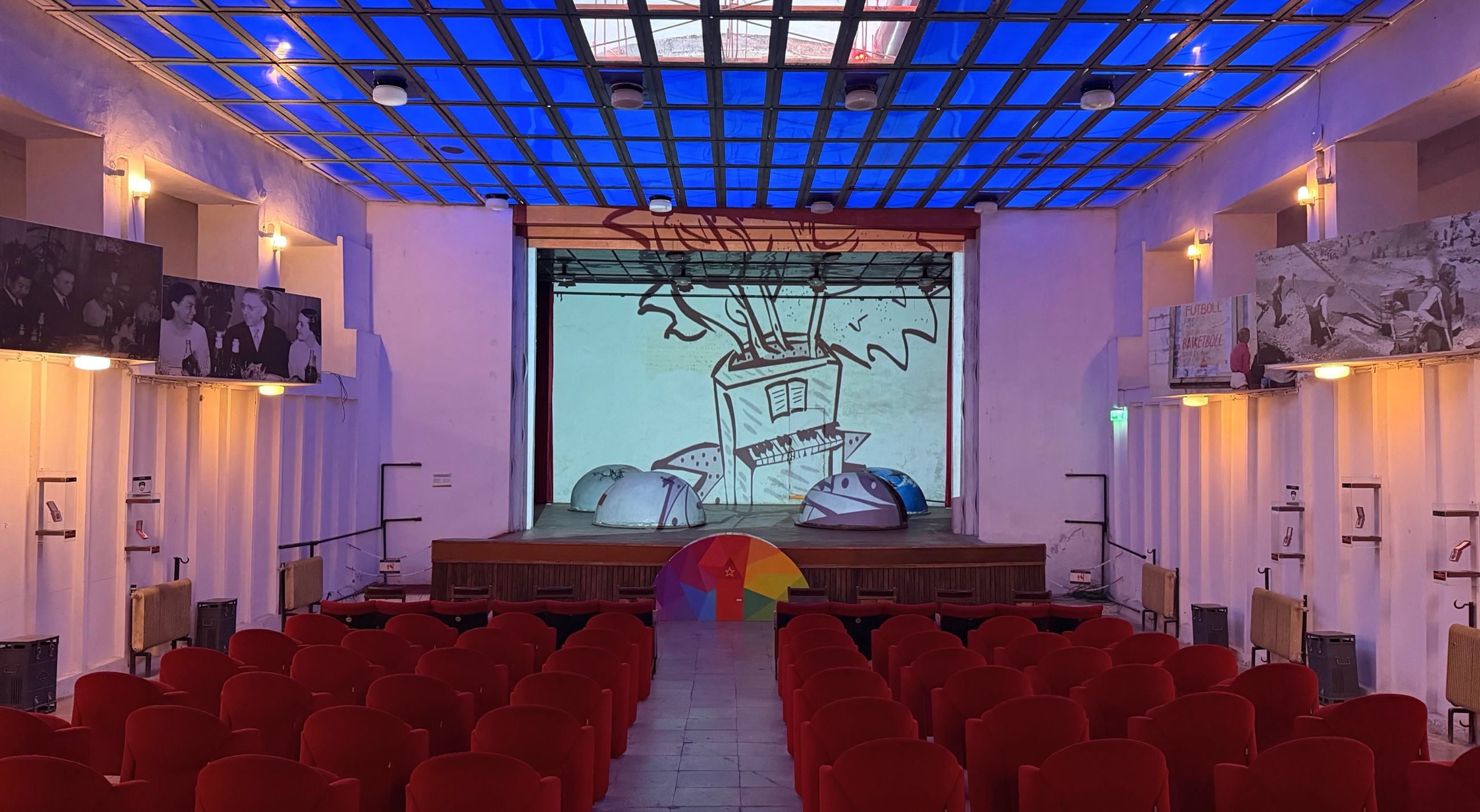 Bunk'Art 1's underground assembly hall with rows of red seats facing a stage beneath a blue-lit ceiling.