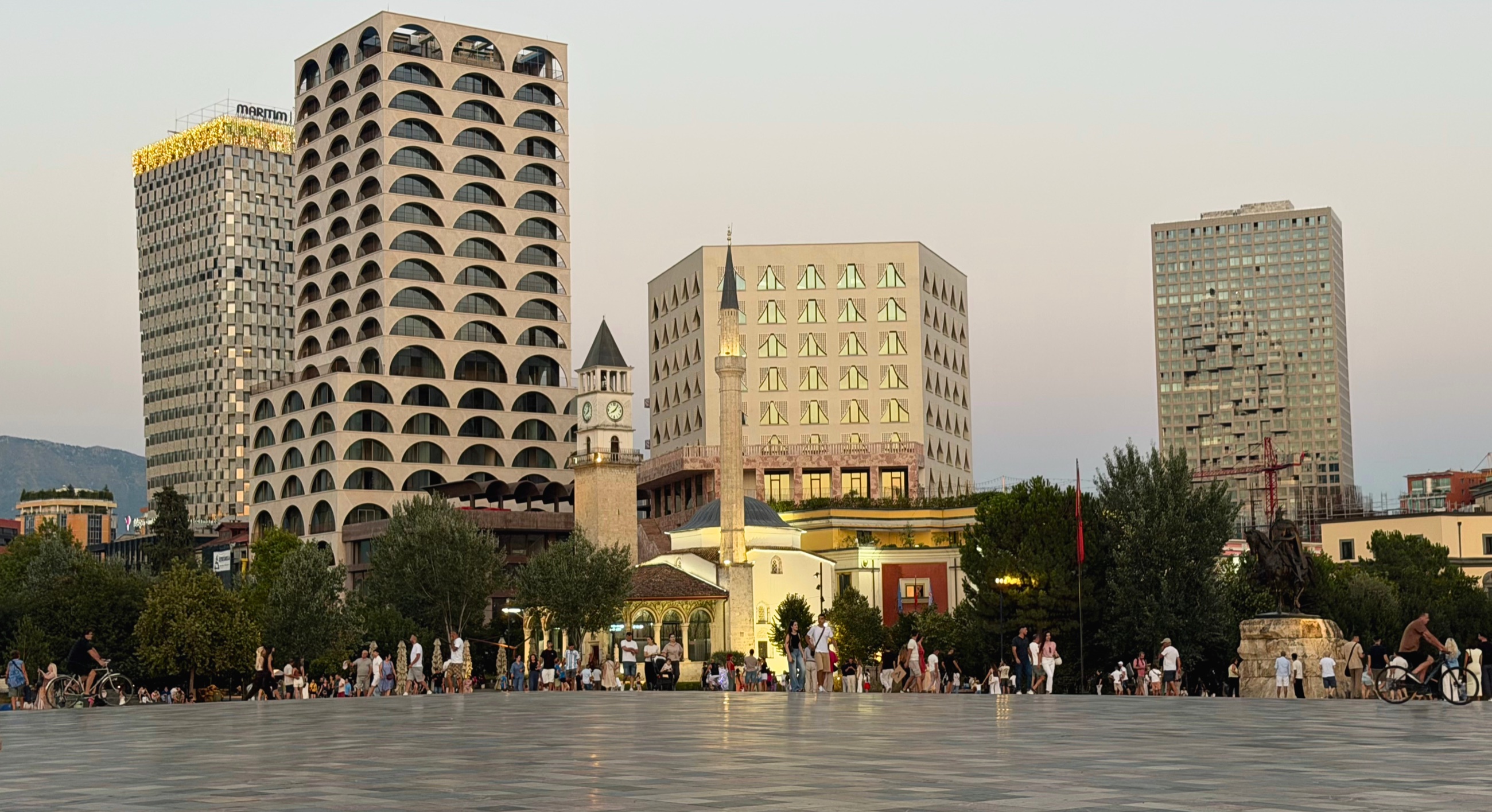Skanderbeg Square at dusk, with the Et'hem Bey Mosque, clock tower, and modern towers surrounding the plaza.