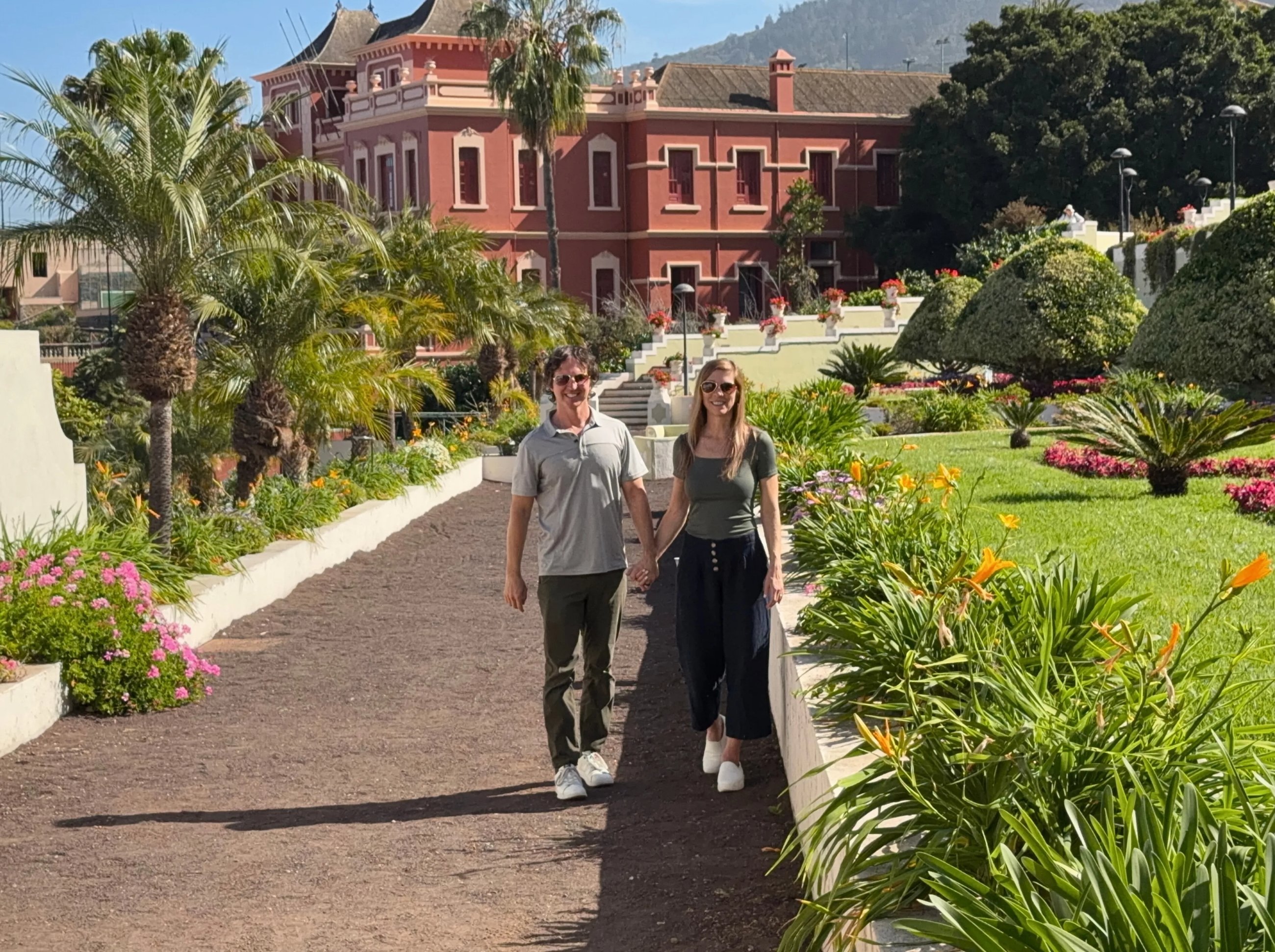 Sam and Stephanie walking hand in hand along a garden path, with a red colonial-era building and mountains behind them.